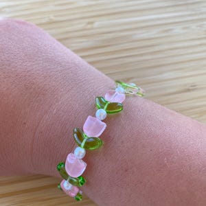 May include: A beaded bracelet featuring pink tulip-shaped beads, green leaf-shaped beads, and small white beads. The bracelet is strung on a clear elastic cord. The bracelet is worn on a wrist.