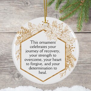 May include: White ceramic Christmas ornament with gold floral design and text. The text reads: "This ornament celebrates your journey of recovery, your strength to overcome, your heart to forgive, and your determination to heal."