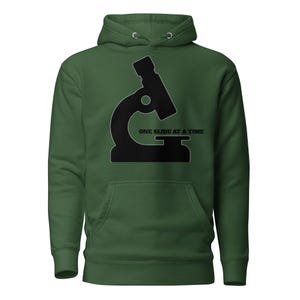 May include: A green hoodie with a black graphic of a microscope and the text "ONE SLIDE AT A TIME". The hoodie has a front pocket and a hood with drawstrings. The design is centered on the front of the garment.