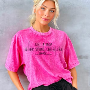 Könnte beinhalten: Ein leuchtend pinkfarbenes T-Shirt mit dem schwarzen Schriftzug "JUST A MOM IN HER STRING CHEESE ERA". Das Shirt hat einen leicht verwaschenen, Vintage-Look und eine lockere Passform. Eine schwarze Schleife befindet sich unter dem Text.