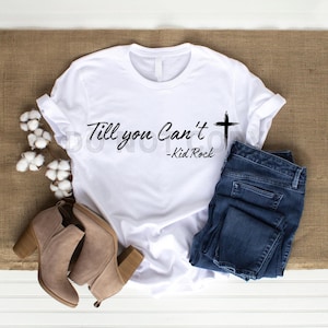 May include: White t-shirt with the text "Till you Can't" and a cross graphic, with "-Kid Rock" below. The shirt is paired with blue jeans and beige ankle boots. The items are arranged on a neutral surface.