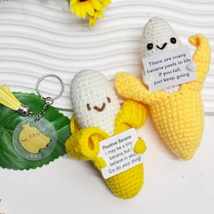 Könnte beinhalten: Zwei gehäkelte Bananen-Schlüsselanhänger mit süßen Gesichtern. Ein Schlüsselanhänger hat eine gelbe Schale und der andere eine weiße Schale. Beide Schlüsselanhänger haben inspirierende Botschaften daran befestigt. Die Botschaft auf dem gelben Bananen-Schlüsselanhänger lautet "Es gibt viele Bananenschalen im Leben. Wenn du fällst, mach einfach weiter." Die Botschaft auf dem weißen Bananen-Schlüsselanhänger lautet "Positive Banane. Ich bin vielleicht eine kleine Banane, aber ich glaube an dich. Mach dein Ding!"