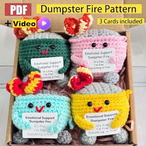 Könnte beinhalten: Ein Set aus vier gehäkelten Mülltonnen-Feuer-Spielzeugen in einem Karton. Jedes Spielzeug hat eine andere Farbe: grün, rosa, blau und gelb. Jedes hat einen grauen Deckel, schwarze Augen und ein rotes Lächeln. Jedes hält ein Schild mit dem Text "Emotional Support Dumpster Fire".