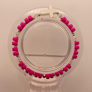 May include: A bright pink beaded bracelet with a white cross charm. The bracelet features small, round pink beads, interspersed with clear crystal beads and gold-colored accents. The cross is centered on the bracelet.