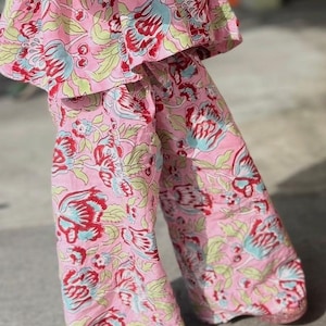 May include: A two-piece outfit consisting of a short-sleeved top and wide-leg trousers, both featuring a light pink base colour with a repeating floral pattern in shades of dark red, light blue, and pale green throughout the fabric.