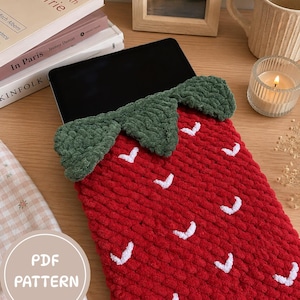 May include: A red crocheted tablet sleeve shaped like a strawberry with a green crocheted leaf top and small white V-shaped accents. The sleeve holds a black tablet and features a circular overlay with the text PDF PATTERN in white letters.