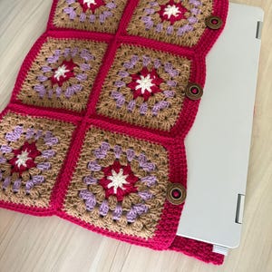 May include: A handmade crochet laptop sleeve featuring a granny square design. The sleeve is made with brown, purple, white, and fuchsia yarn. It has a fuchsia border and wooden button closures. The sleeve is designed to fit a laptop.