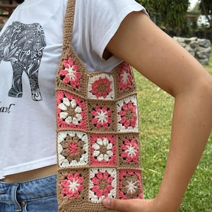 Puede incluir: Bolso tote de crochet hecho a mano con un patrón floral en tonos rosa, blanco y marrón. El bolso tiene forma rectangular con borde y correa marrones. El bolso lo sostiene una persona con camiseta blanca y vaqueros. Medidas: 30 cm x 30 cm.