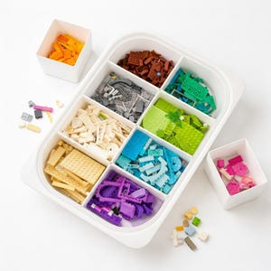 Puede incluir: Un organizador de plástico blanco lleno de bloques de construcción de juguete coloridos. Los bloques están clasificados por color, incluyendo amarillo, marrón, verde, azul y morado. Dos contenedores blancos adicionales contienen más bloques. Se ven varias formas y tamaños.