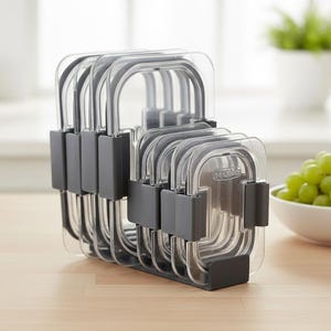 Puede incluir: Un juego de recipientes rectangulares transparentes para guardar alimentos con asas y tapas grises, apilados ordenadamente en un soporte gris. Los recipientes están sobre una superficie de madera clara. Un cuenco de uvas verdes está en el fondo.