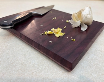 May include: A purple wooden cutting board with a silver knife and a clove of garlic that has been peeled and minced. The garlic is scattered on the board.