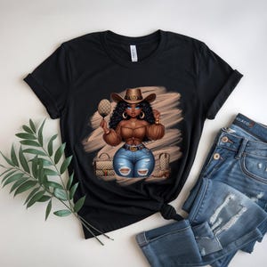 May include: Black t-shirt featuring a cartoon illustration of a woman in a cowboy hat, brown top, and blue jeans. The design includes a purse, boots, and a mirror. The shirt is paired with blue jeans.