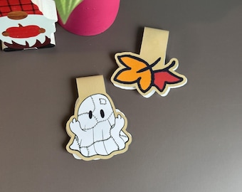 Puede incluir: Dos marcapáginas magnéticos con diseños planos troquelados. Uno muestra un fantasma blanco con detalles de costuras negras y un parche. El segundo marcapáginas muestra dos hojas de otoño superpuestas en naranja y rojo con contornos negros.