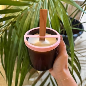 Può includere: Un barattolo di candela in vetro marrone con un bordo rosa e un supporto per stoppino in legno. La candela ha un interno in cera bianca. Il barattolo è tenuto davanti a una pianta verde fogliosa.