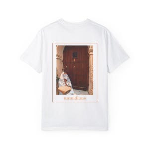 May include: White t-shirt with a photo of a person in a white robe standing in front of a wooden door. The text "numidians" is printed below the photo.