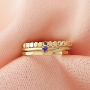 May include: Three gold rings stacked on top of each other. The top ring is a band of small gold circles. The middle ring is a thin, plain band. The bottom ring has a small purple gemstone in the centre.