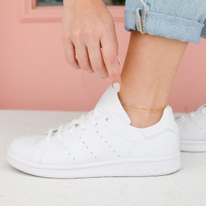 May include: A gold-colored chain anklet is worn over a white sneaker. The anklet has two delicate strands with a link design. The sneaker is white with the Adidas logo on the side. The person is wearing blue jeans.