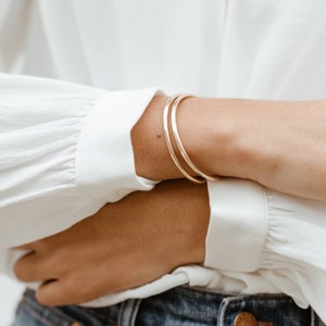 May include: Two gold bangle bracelets stacked on a person's wrist. The bracelets are simple and minimalist in design.