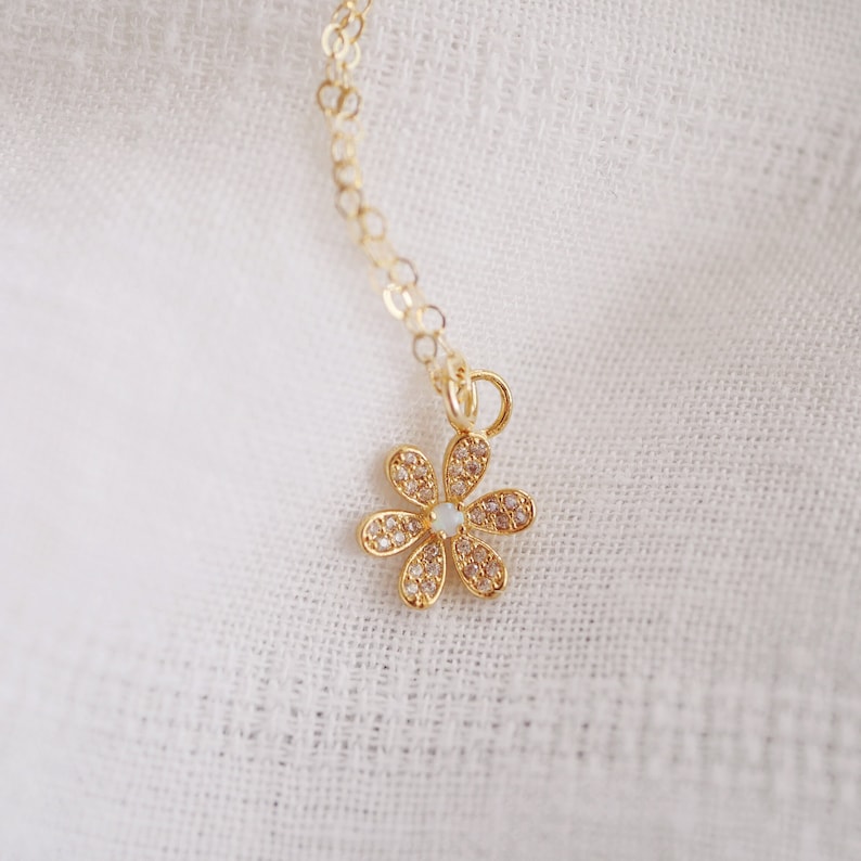 Peut inclure: Un collier en cha&icirc;ne dor&eacute;e avec un petit pendentif en forme de fleur scintillante. La fleur a un centre blanc et est entour&eacute;e de strass transparents.