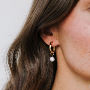 Op de afbeelding: Gouden creolen met een hangende parelbedel. De oorbellen worden gedragen door een persoon met lang bruin haar.