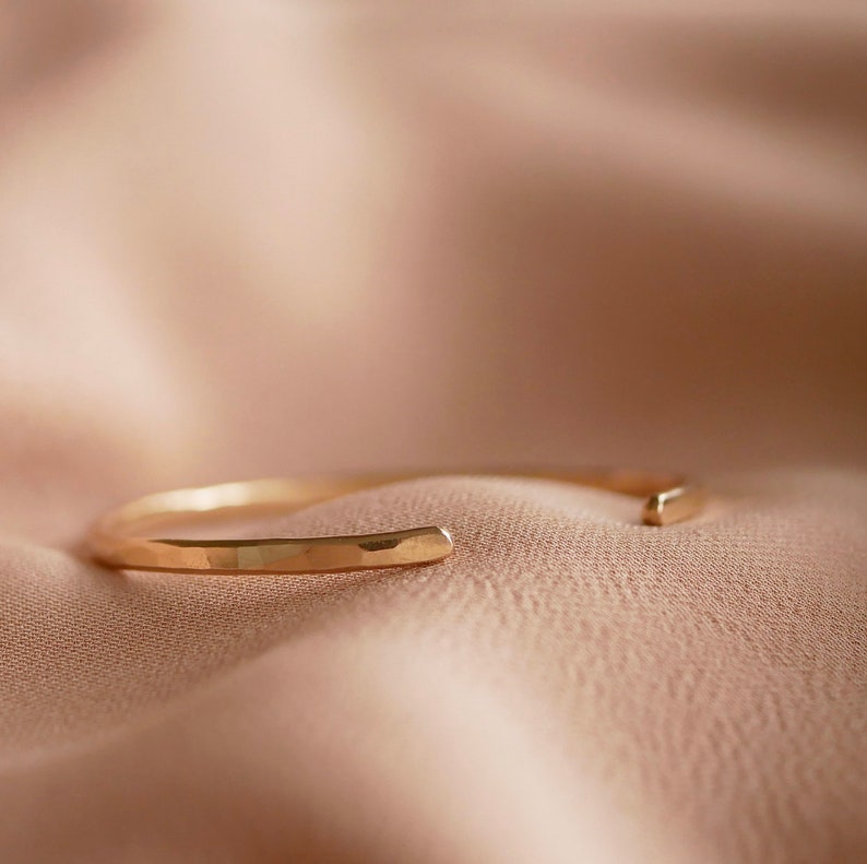May include: A simple gold bracelet with a hammered finish. The bracelet is open and has a flat, rectangular shape.