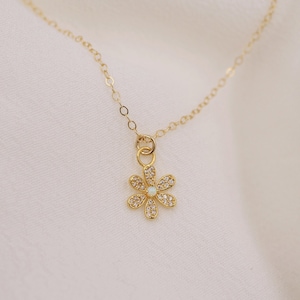 Peut inclure: Un collier en cha&icirc;ne dor&eacute;e avec un petit pendentif en forme de fleur scintillante. La fleur a un centre en opale blanche et est entour&eacute;e de pierres de zircone cubique transparentes.