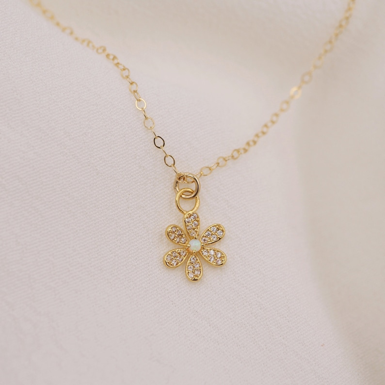Peut inclure: Un collier en cha&icirc;ne dor&eacute;e avec un petit pendentif en forme de fleur scintillante. La fleur a un centre en opale blanche et est entour&eacute;e de pierres de zircone cubique transparentes.