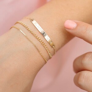 May include: A gold chain bracelet with a thin chain, a thicker chain, and a bar charm with the inscription "91.48W 30.2472N".