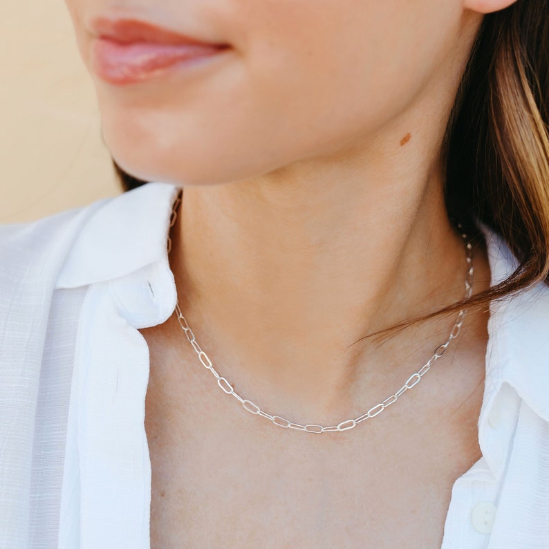 May include: A silver chain necklace with a delicate, oval-shaped link design. The necklace is worn by a person with a white shirt.