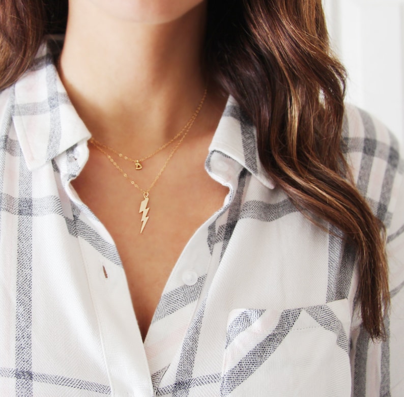 May include: A gold necklace with a lightning bolt pendant and a small charm. The necklace is layered on a person wearing a white and gray plaid shirt.