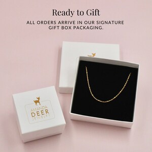 K&ouml;nnte beinhalten: Zwei wei&szlig;e Geschenkboxen mit schwarzem Innenraum. Eine Box enth&auml;lt eine goldene Kettenkette. Die andere Box zeigt ein goldenes Hirschlogo mit dem Text "Amanda Deer Jewelry".