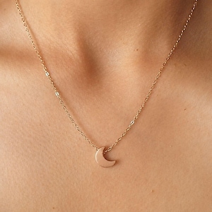 May include: A gold-toned necklace featuring a delicate chain and a small, smooth crescent moon pendant. The pendant is centered on the chain, which consists of thin, interconnected links that rest against the skin. The metal has a polished finish.