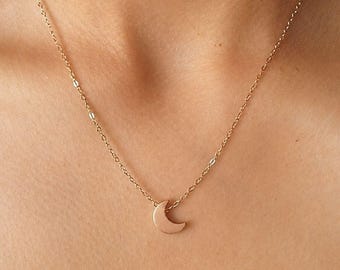 Puede incluir: Un collar de tono dorado que presenta una cadena delicada y un pequeño colgante liso en forma de media luna. El colgante está centrado en la cadena, la cual consiste en eslabones finos e interconectados que descansan sobre la piel. El metal tiene un acabado pulido.