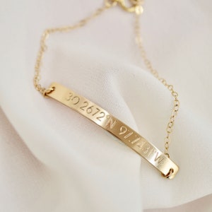 May include: Gold chain bracelet with a thin, curved bar charm engraved with the coordinates "30.2672&deg; N 97.7431&deg; W".