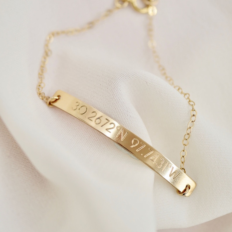 May include: Gold chain bracelet with a thin, curved bar charm engraved with the coordinates "30.2672&deg; N 97.7431&deg; W".