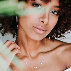 May include: A gold necklace with a crescent moon and three stars charms. The necklace is worn by a person with curly brown hair.