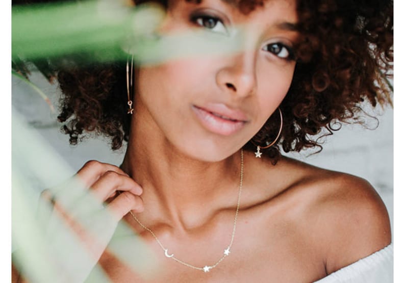 May include: A gold necklace with a crescent moon and three stars charms. The necklace is worn by a person with curly brown hair.