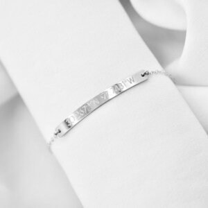 May include: A silver bracelet with a thin chain and a flat bar charm engraved with the coordinates "30.2672&deg;N 97.7431&deg;W".