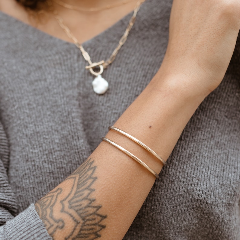 May include: A gold double bangle bracelet on a person's arm. The bracelet is made of two thin bands of gold that are stacked on top of each other.