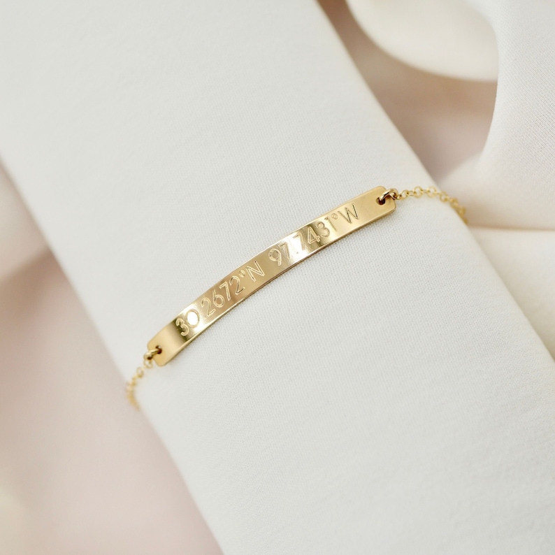 May include: A gold chain bracelet with a thin, flat bar charm engraved with the coordinates "30.2672&deg;N 97.7431&deg;W".