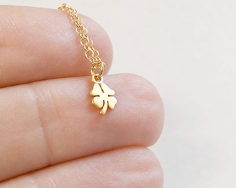 May include: A gold-colored necklace featuring a delicate chain and a small, four-leaf clover pendant. The pendant is suspended from a circular jump ring attached to the fine link chain, showcasing a polished metallic finish throughout the piece.
