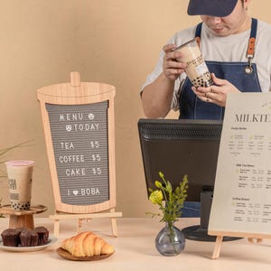 May include: A wooden sign with a letter board displaying a menu with prices for tea, coffee, cake, and boba. A person is holding a clear plastic cup with boba tea. A second sign displays a milk tea menu with prices. There are also pastries and a cup of boba tea on a table.