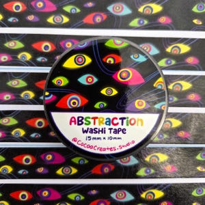May include: A round roll of washi tape featuring a black background with colorful, multi-layered eye patterns in yellow, red, blue, and green. The label reads ABSTRACTION WASHI TAPE 15mm x 10m and @CocoaCreates.Studio in colorful lettering.