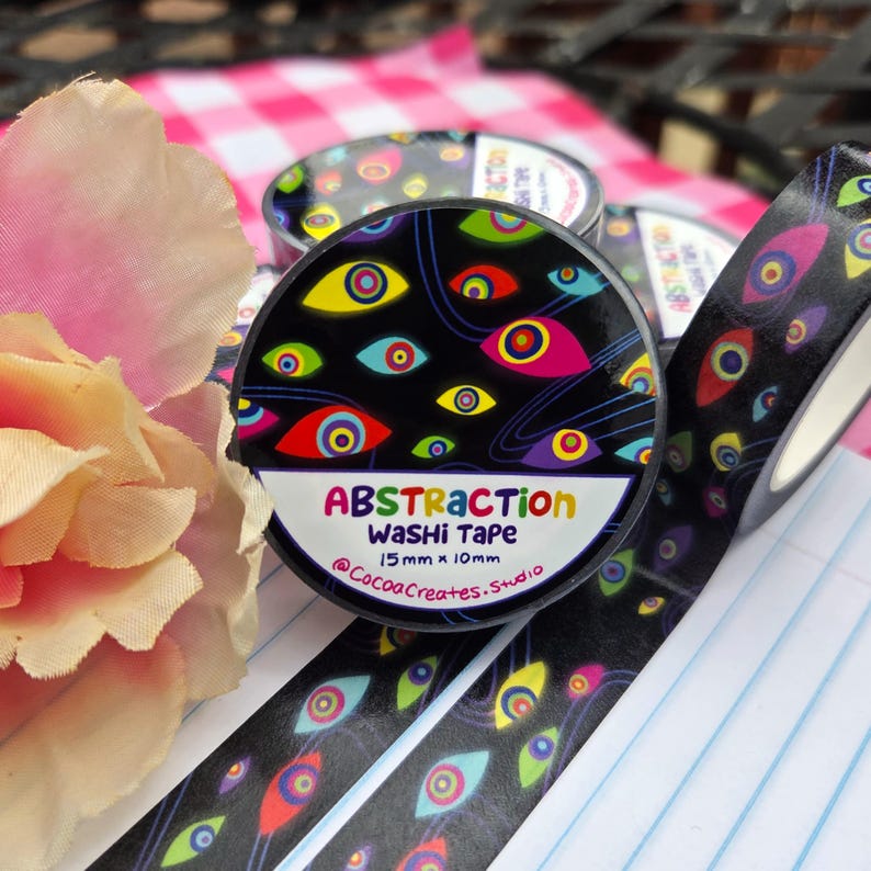 May include: A roll of black washi tape featuring a colorful pattern of stylized eyes in shades of red, yellow, blue, and purple. The label reads Abstraction Washi Tape 15mm x 10mm and @CocoaCreates.Studio. The tape is displayed with a light pink flower.