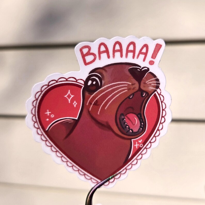 May include: A sticker featuring a cartoon seal with its mouth open, inside a red heart with a scalloped edge. The word "BAAAA!" is written in red above the seal. The sticker has a white border and is held by a metal clip.