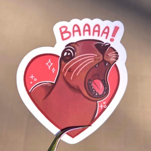 May include: A sticker featuring a cartoon otter with its mouth open, inside a red heart. The otter is brown with white whiskers and the word "BAAAA!" in white text above it. The heart has decorative elements.