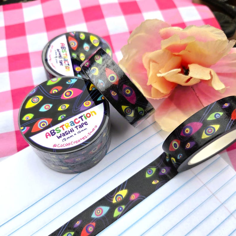 May include: Two rolls of black washi tape featuring a colorful eye pattern with yellow, pink, and blue irises. The label reads Abstraction Washi Tape 15mm x 10m @CocoaCreates.Studio. One roll is partially unrolled to show the repeating eye design.