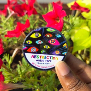 May include: A circular roll of washi tape featuring a black background with colorful, stylized eye patterns. The label displays the text ABSTRACTION Washi Tape, 15mm x 10mm, and @CocoaCreates.studio in a white, semi-circular section at the bottom.