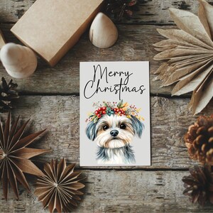 K&ouml;nnte beinhalten: Eine Weihnachtskarte mit einer Aquarell-Illustration eines kleinen Hundes, der einen Kranz aus Blumen und Beeren tr&auml;gt. Die Karte tr&auml;gt den Text "Merry Christmas" in schwarzen Buchstaben.