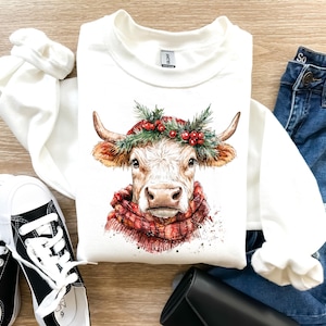 K&ouml;nnte beinhalten: Wei&szlig;er Pullover mit einer Aquarell-Illustration einer Kuh, die einen roten und gr&uuml;nen Kranz mit Beeren tr&auml;gt. Die Kuh hat einen rot-wei&szlig;en Schal um den Hals.